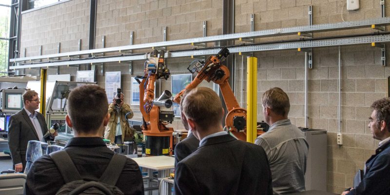 Zwei Roboterarme, die gemeinsam ein WerkstÃ¼ck bearbeiten, werden vor Zuschauern bei der Auftaktveranstaltung prÃ¤sentiert.
